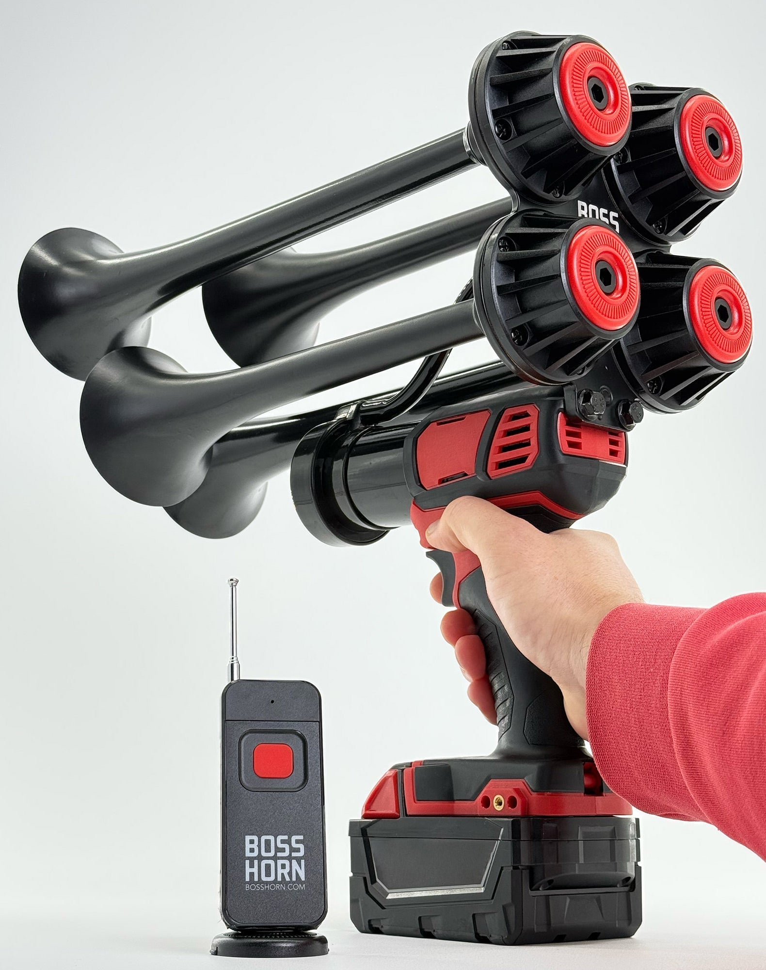 Hand holding the black quad-trumpet horn with red caps on a Milwaukee® M18 battery, antenna remote beside it on a light backdrop