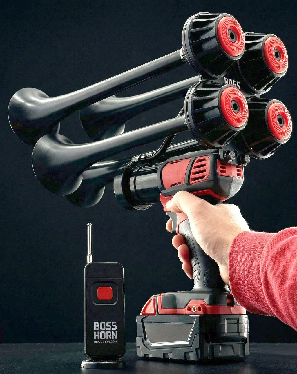 Person aiming the black-and-red four-trumpet train horn upward by the handle, remote and battery in foreground on a dark background