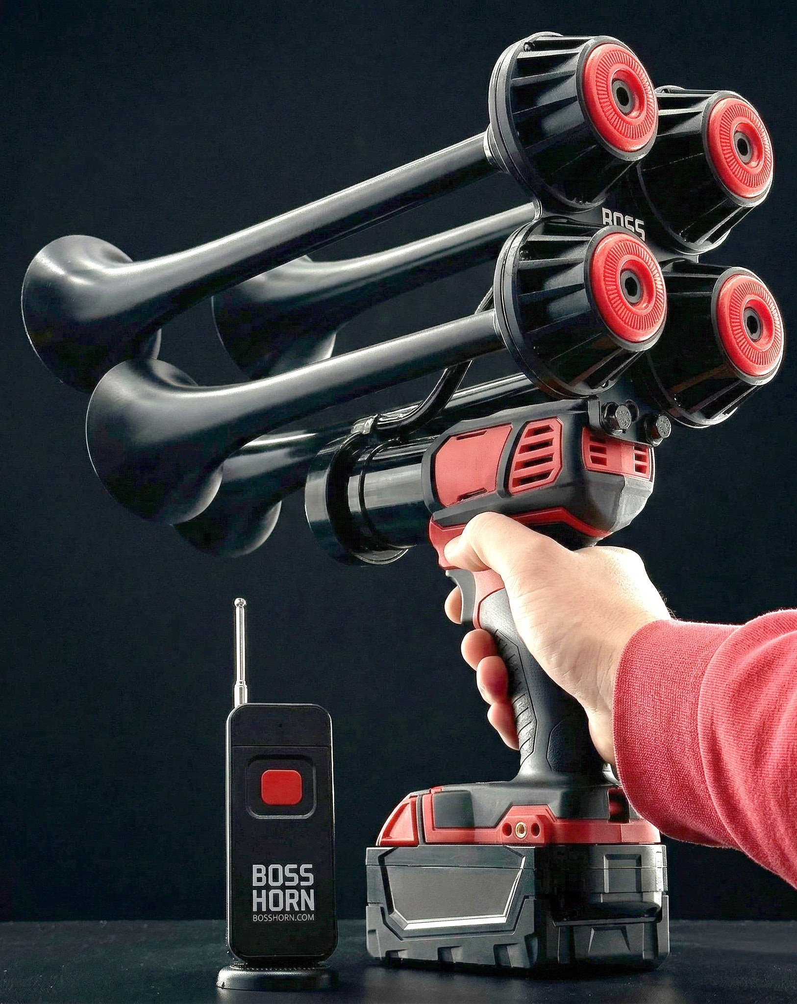 Person aiming the black-and-red four-trumpet train horn upward by the handle, remote and battery in foreground on a dark background