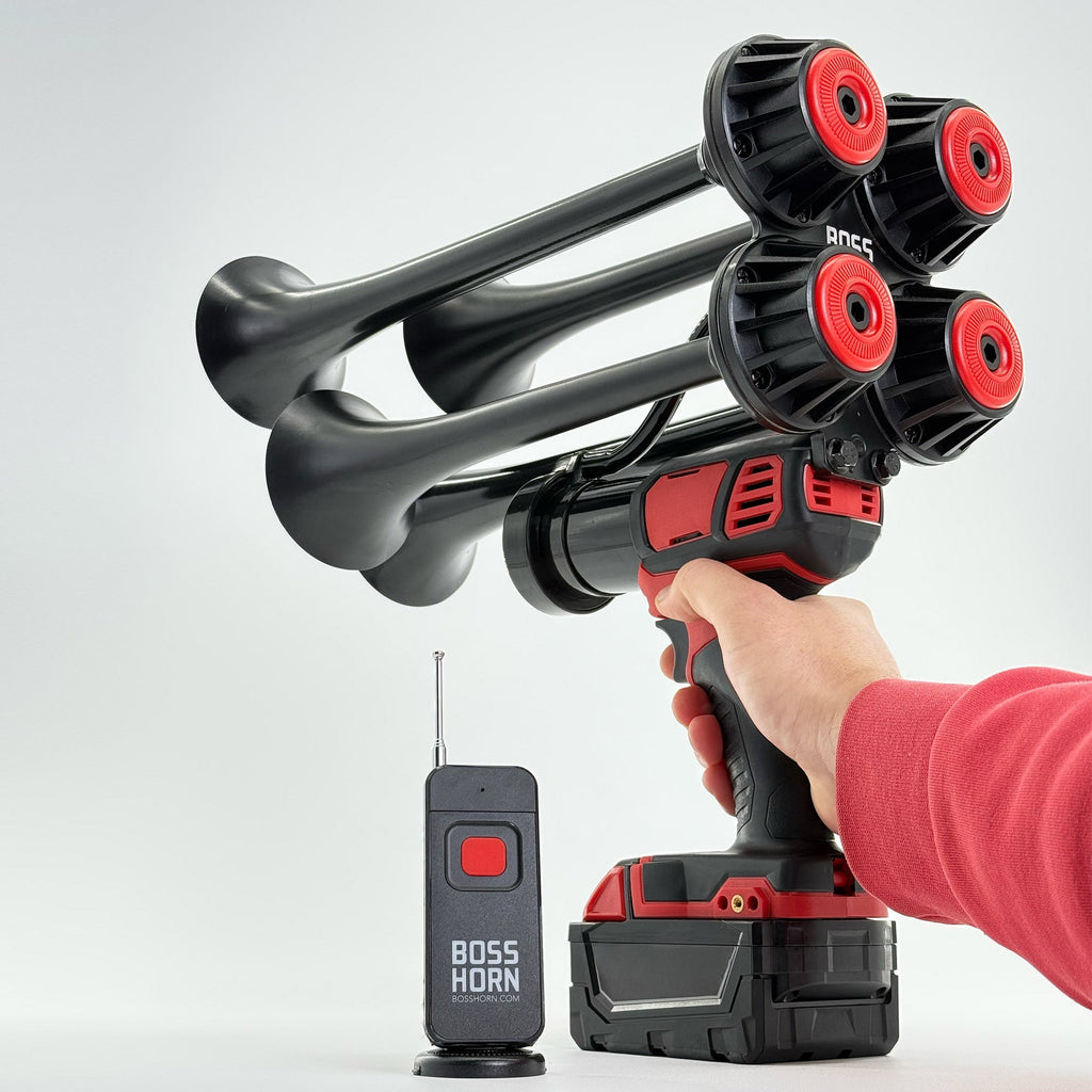 Hand holding the four-trumpet horn powered by a Milwaukee® M18 battery, wireless antenna remote beside it on a light background