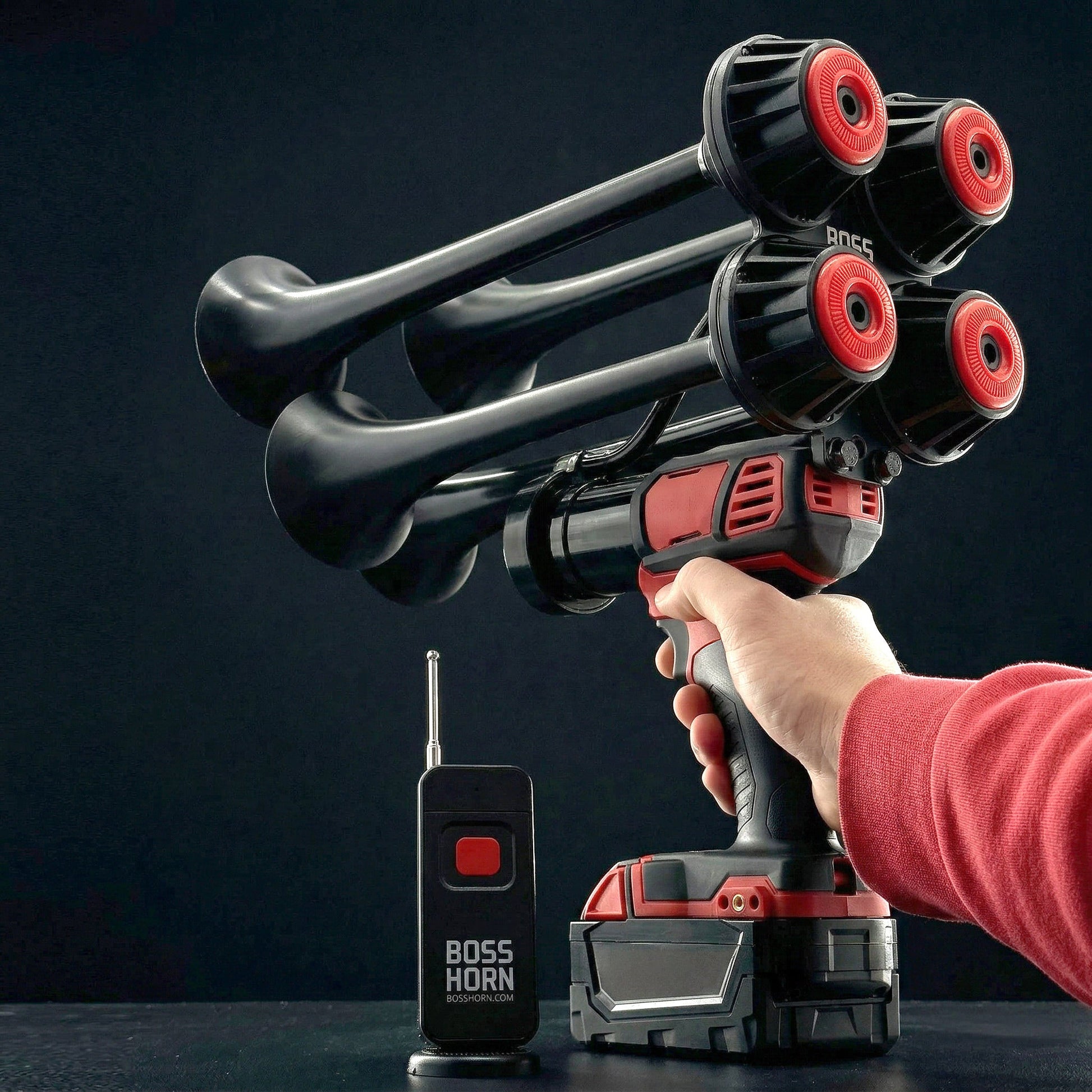 Hand gripping the train horn by its trigger handle, aiming the black-and-red quad trumpets upward, with remote and battery on a dark surface
