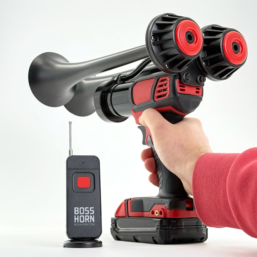 Hand holding and aiming the black dual-trumpet train horn on a red cordless power tool with Milwaukee® M18 battery, remote nearby on white